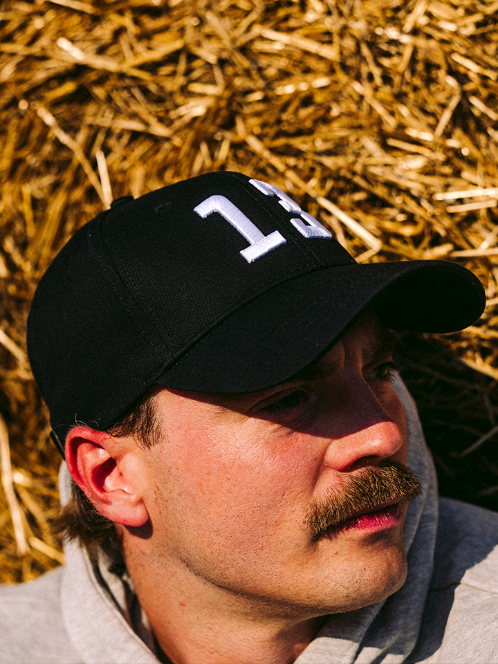 A mustached person in a light grey hoodie wears the black 13 - Casual Cap by La Machine Cycle Club, lying on straw.