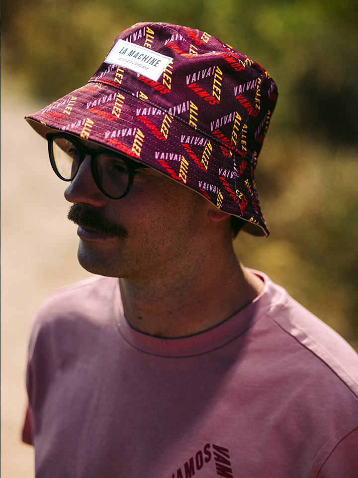 A person in glasses, a thick mustache, a light pink shirt, and a La Machine Cycle Club La Machine - Reversible Bucket Hat with yellow and white “VAIVA” text and a “LA MACHINE” patch stands outside in the sunlight.