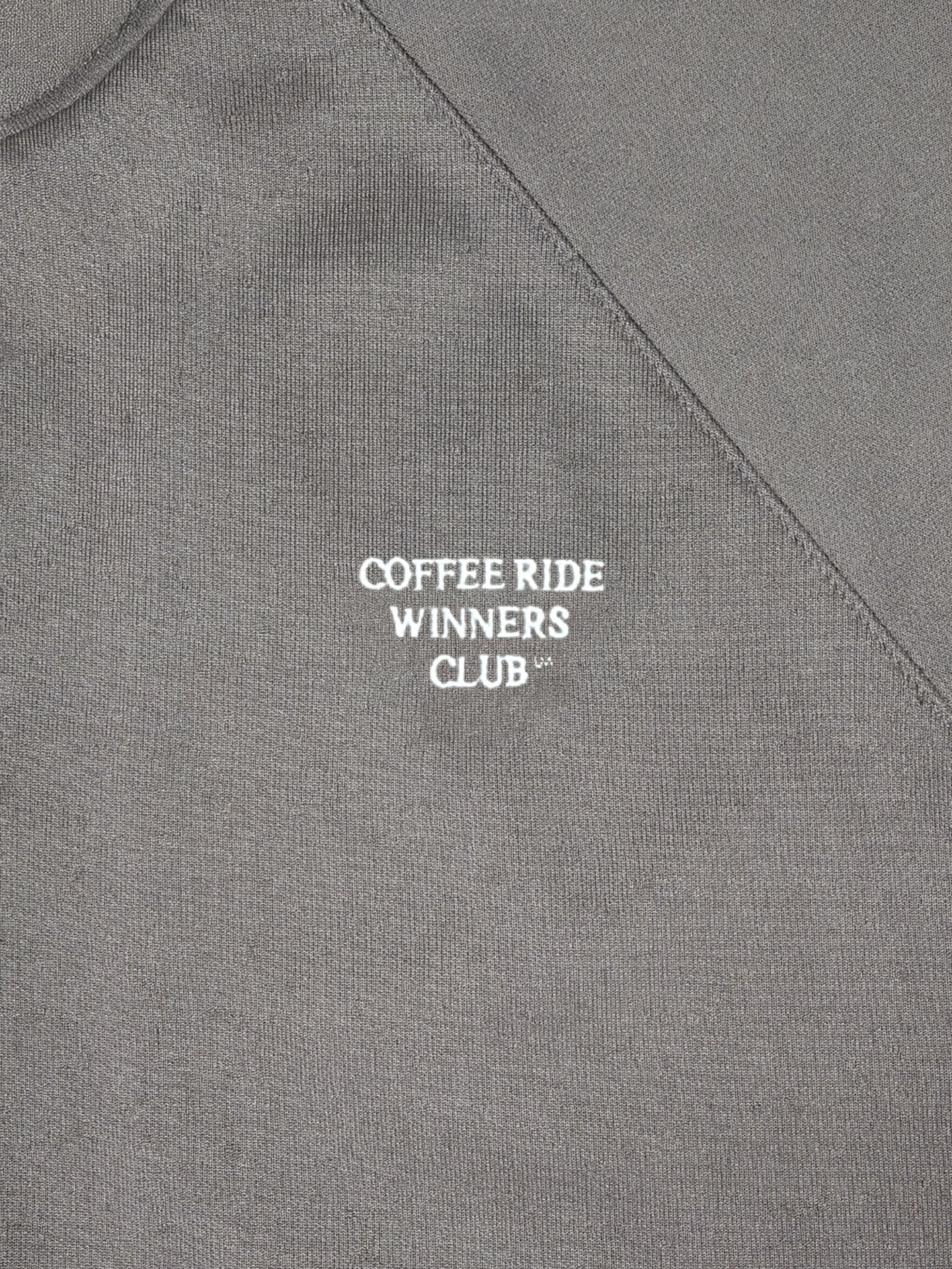 Close-up of the Coffee Ride Winners Club - Track Jacket - Steel Grey by La Machine Cycle Club, featuring white COFFEE RIDE WINNERS CLUB™ text on angled gray fabric—ideal for fans of the coffee ride tradition.