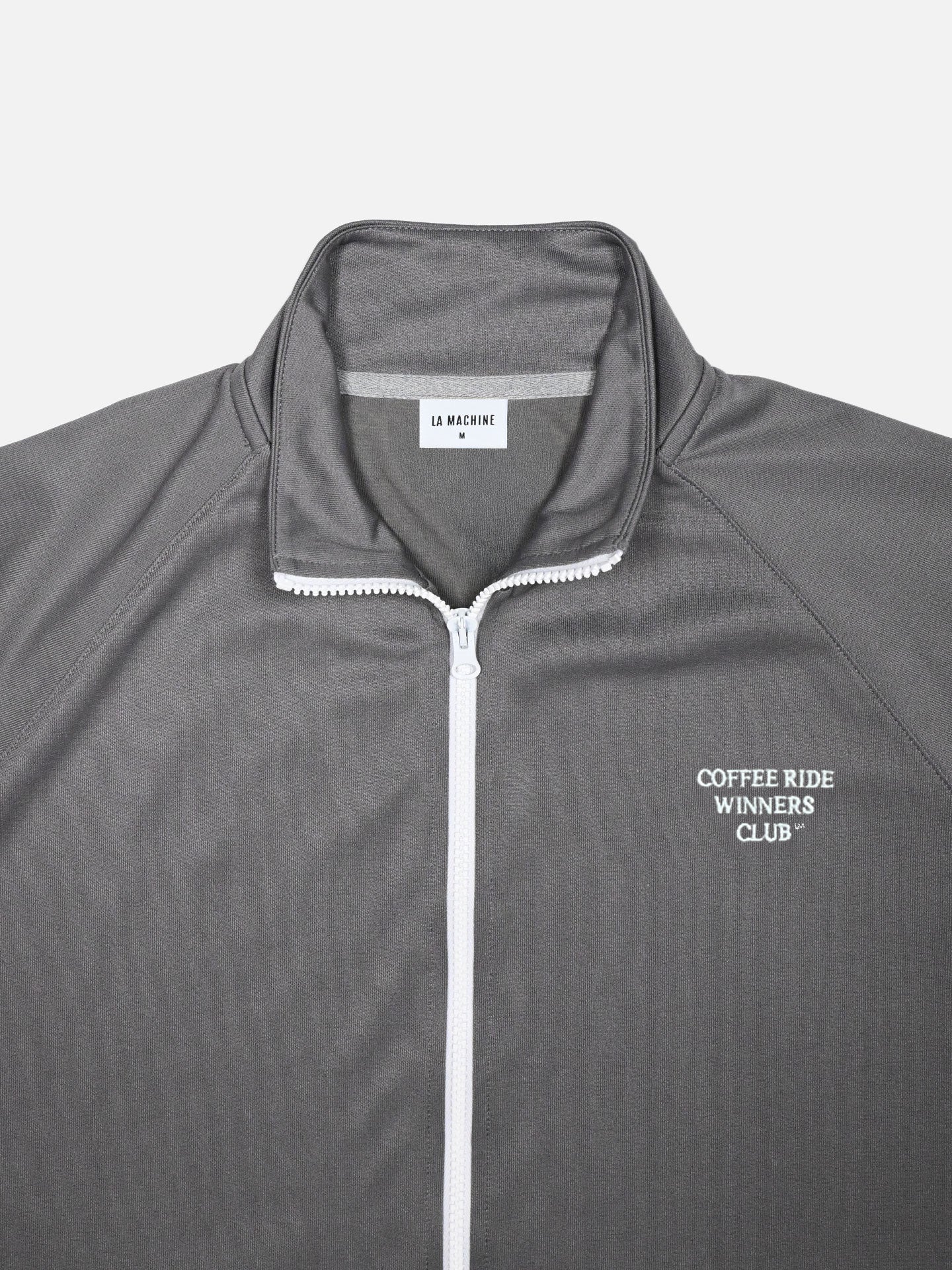 A close-up of the Coffee Ride Winners Club - Track Jacket in steel grey by La Machine Cycle Club, featuring a white zipper, COFFEE RIDE WINNERS CLUB® text on the chest, and a collar label with the brand name.