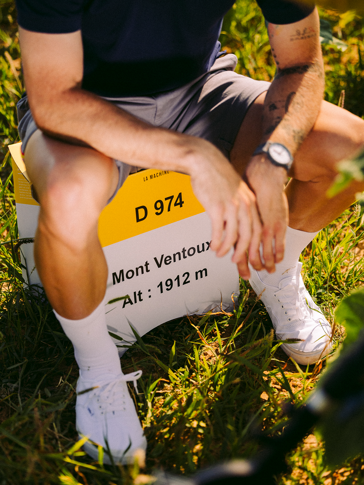 A person in shorts and white sneakers sits on a La Machine Cycle Club Mont Ventoux - 2-in-1 Stool & Sidetable, styled as a D 974 Mont Ventoux Alt: 1912 m milestone marker, surrounded by grass. Their face is not visible.