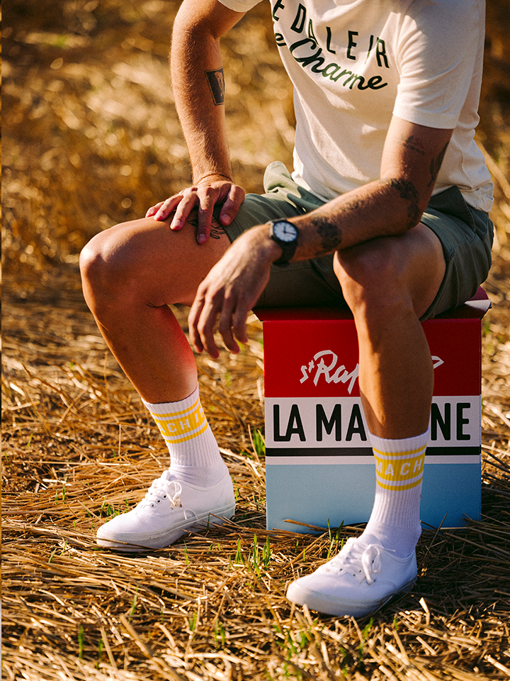 A person in vintage racing style sits relaxed on the St. Raphaël 2-in-1 Stool & Sidetable by La Machine Cycle Club, wearing a white t-shirt, green shorts, and sneakers in a sunlit field.