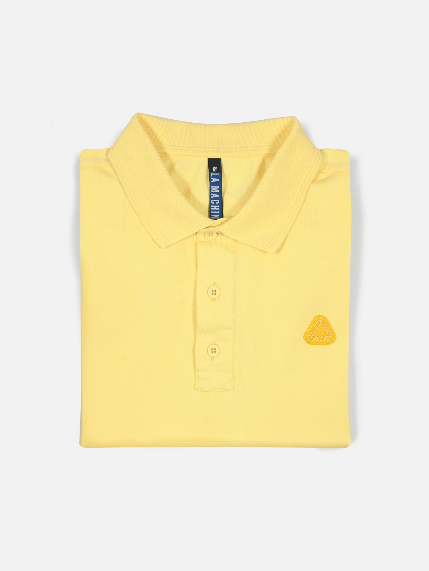 The Allez - Tour de France Polo Shirt by La Machine Cycle Club features a light yellow unisex design with a collar, three buttons, and a small 3D rubber badge on the left chest, displayed against a plain white background.