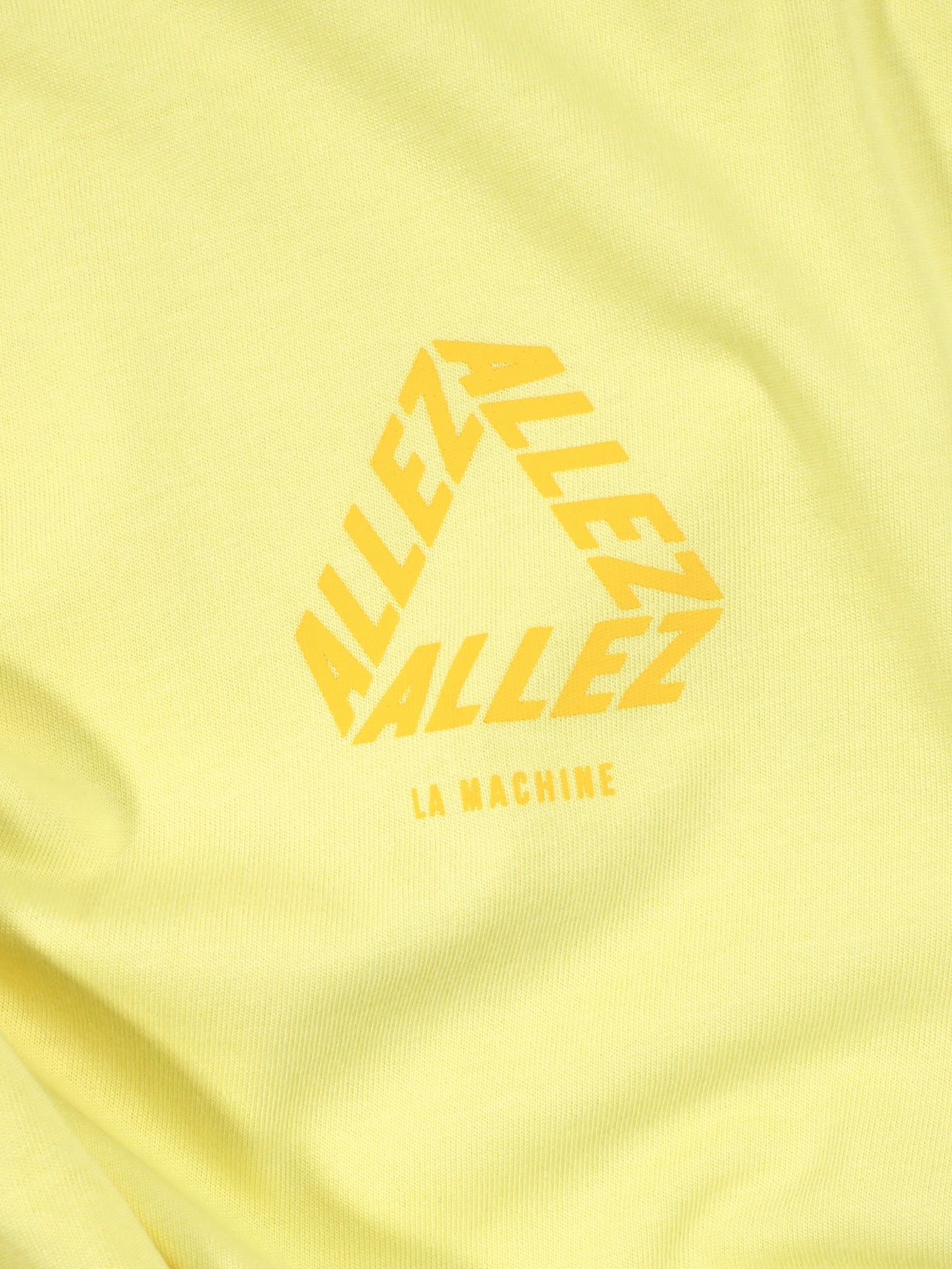 The Allez - Loose Fit - Tour de France T-shirt by La Machine Cycle Club features yellow fabric with a triangle of ALLEZ repeated in orange and LA MACHINE printed below—perfect for cycling enthusiasts.