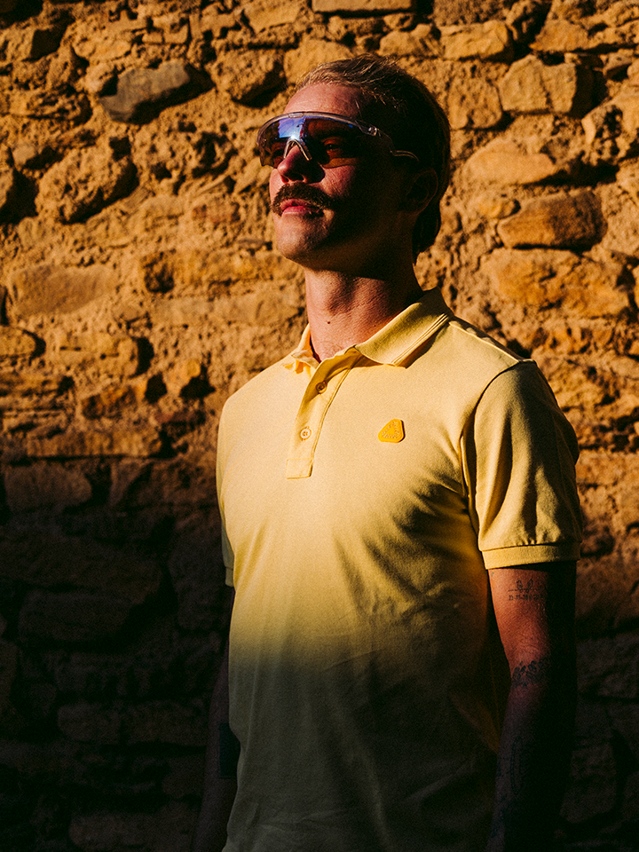 A person in La Machine Cycle Clubs Allez - Tour de France Polo Shirt in light yellow and sunglasses stands against a sunlit stone wall, with sunlight highlighting part of their face and shirt while the rest remains in shadow.