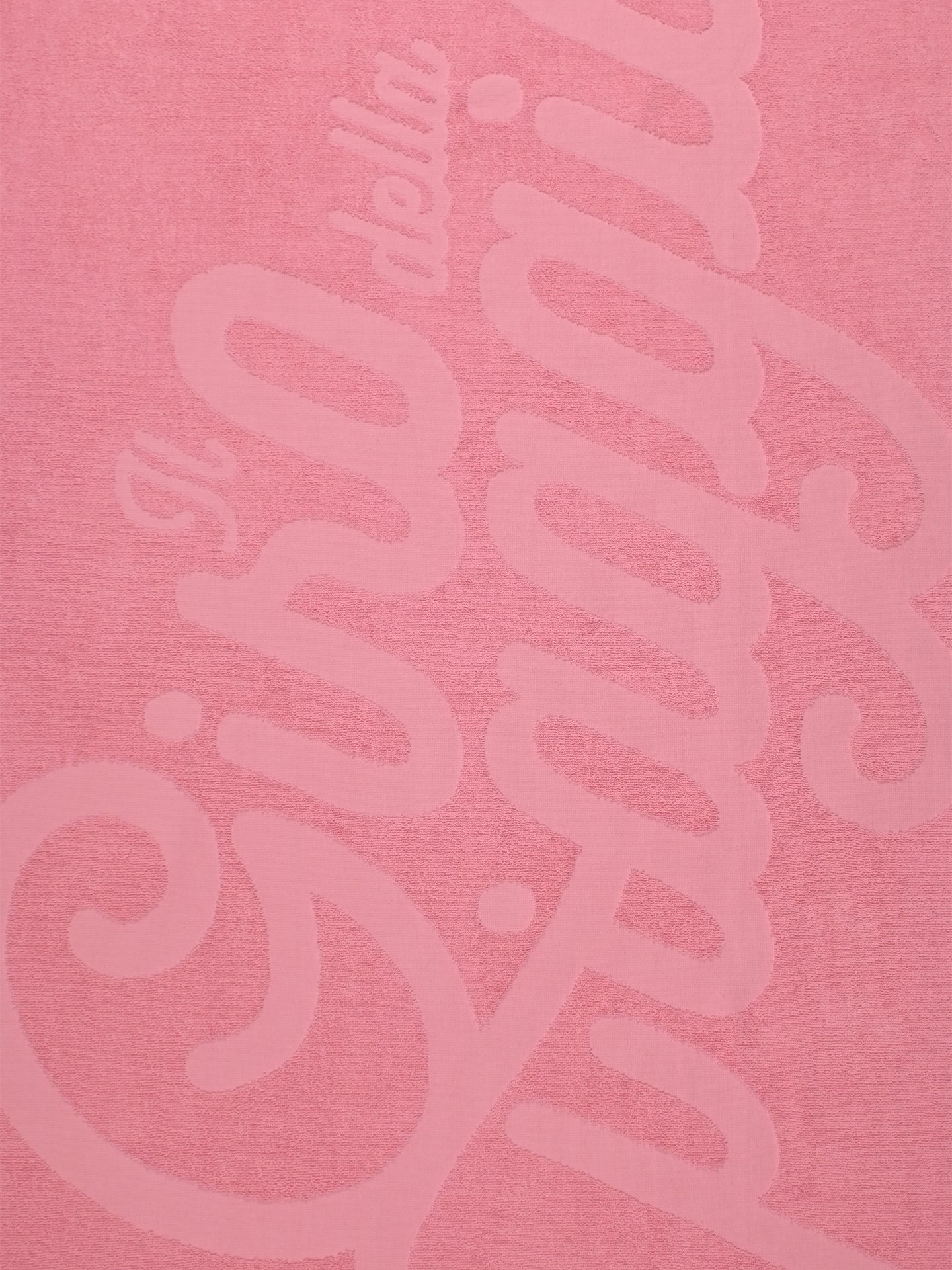 The Il Giro della Spiaggia Beach Towel by La Machine Cycle Club features a pink textured surface with raised, cursive Italian text. Inspired by The Grand Tour, its elegant design contrasts subtly in darker pink on premium cotton.