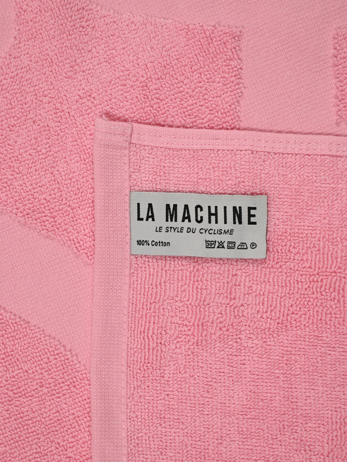A close-up of the Il Giro della Spiaggia beach towel by La Machine Cycle Club in pink, featuring a white label with 100% Cotton, LA MACHINE LE STYLE DU CYCLISME, and wash care symbols.