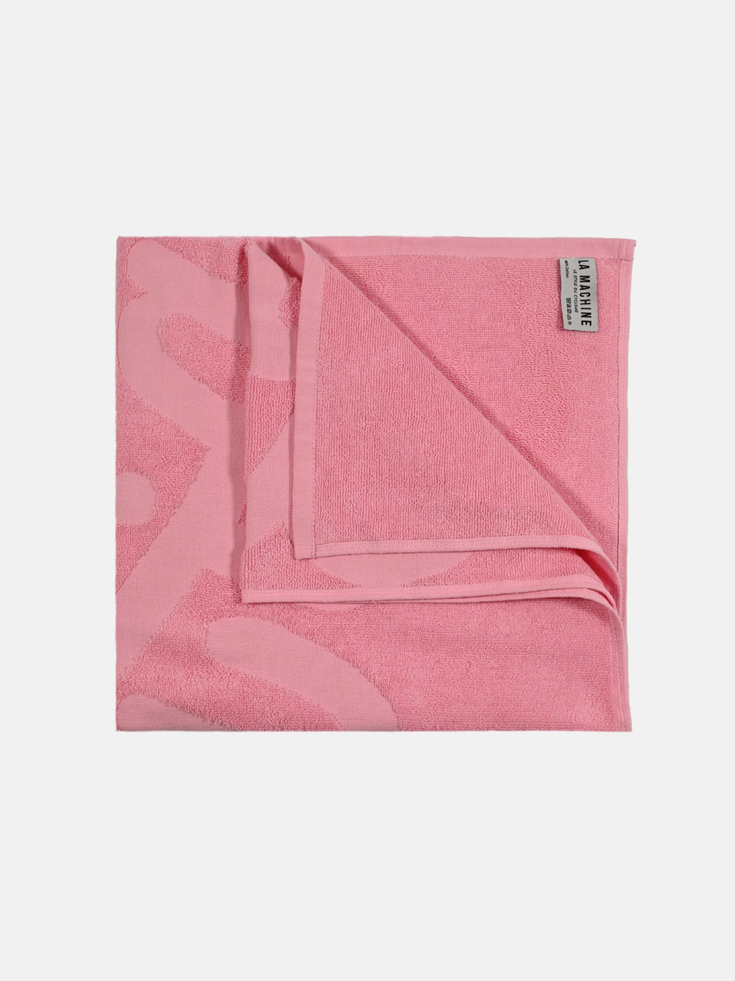 A neatly folded pink Il Giro della Spiaggia Beach Towel by La Machine Cycle Club with a subtle pattern and a rectangular label in the top right corner is displayed on a plain white background.