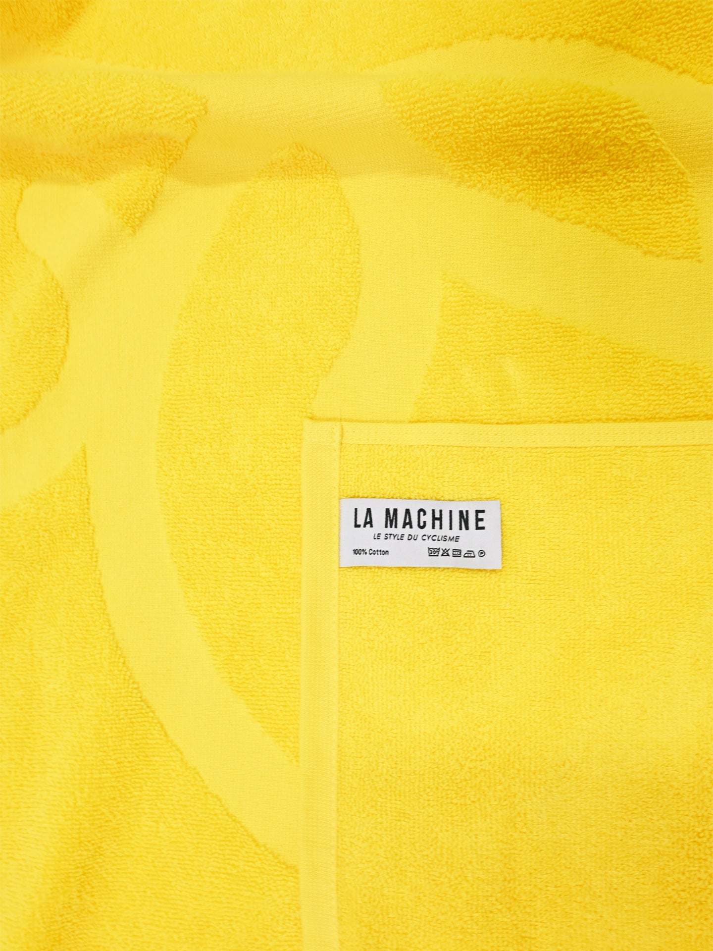A vibrant yellow Le Tour de la Plage beach towel by La Machine Cycle Club, featuring a subtle pattern and a white label with the brand name plus care and material details.