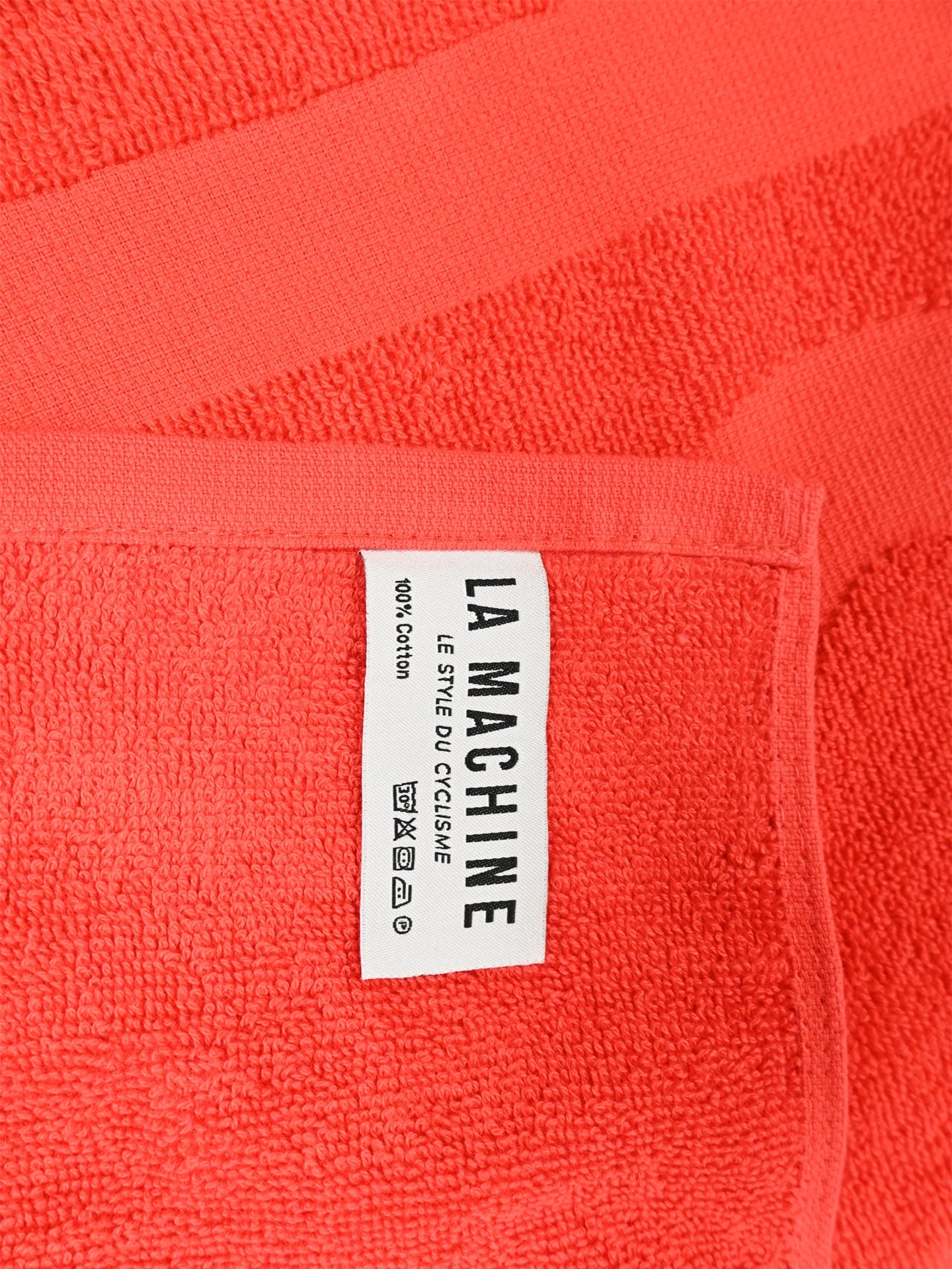 A close-up of the La Machine Cycle Club’s La Vuelta a la Playa - Beach Towel in coral, featuring a stitched white label reading LA MACHINE, LESPRIT DU CYCLISME, 100% COTON with care symbols—perfect for cycling enthusiasts at the beach.