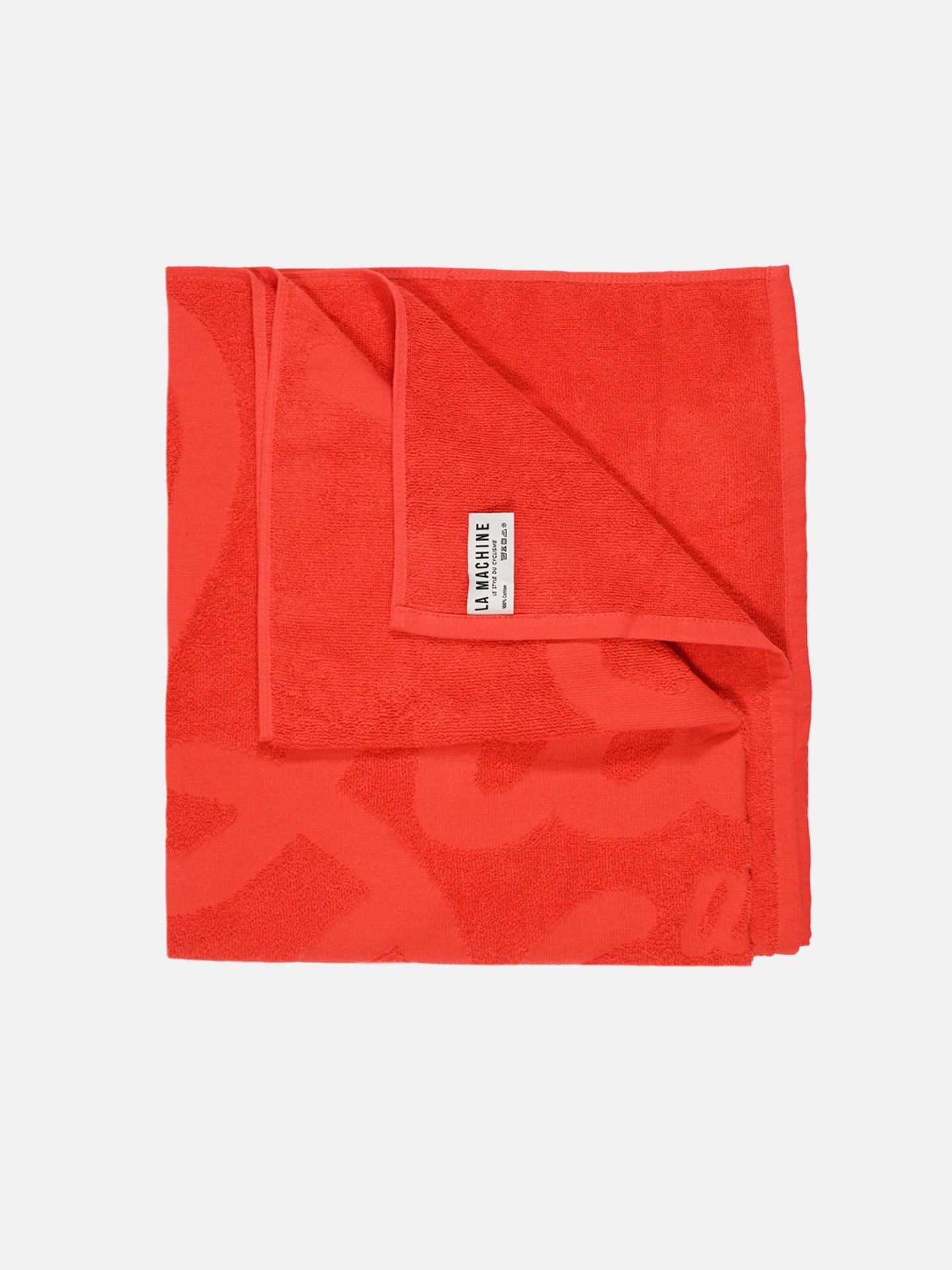 A vivid red La Vuelta a la Playa beach towel by La Machine Cycle Club, made from premium cotton with a subtle texture, is neatly folded. A white label reading “LA MANUFACTURE” appears on the corner. The background is plain white.