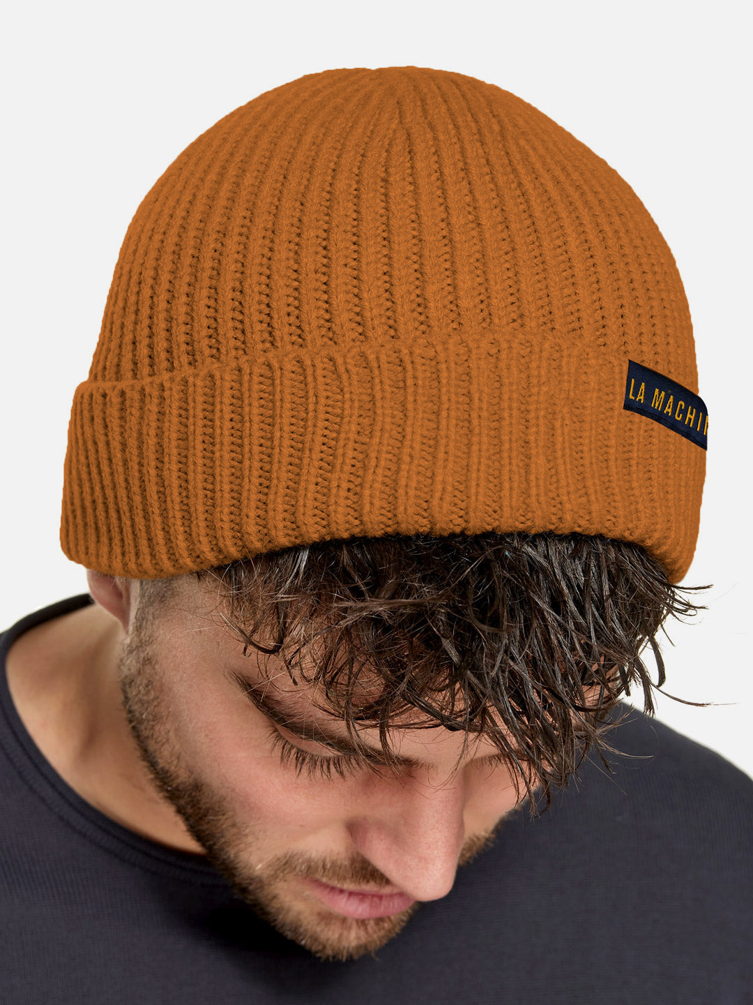 A man with wet, tousled hair looks down wearing the La Machine Cycle Club La Machine - Beanie - Burnt Orange, featuring orange ribbed knit and a black LA MACHINE label, paired with a dark shirt against a plain, light background.