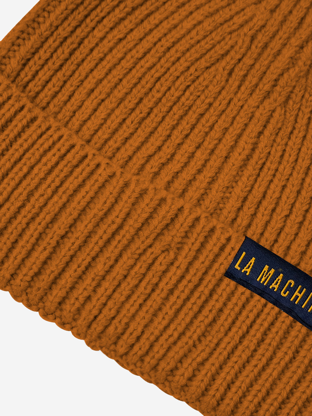 Close-up of the ribbed, burnt orange La Machine - Beanie by La Machine Cycle Club, a unisex winter essential featuring a navy blue label with yellow text partially reading LA MACHI.