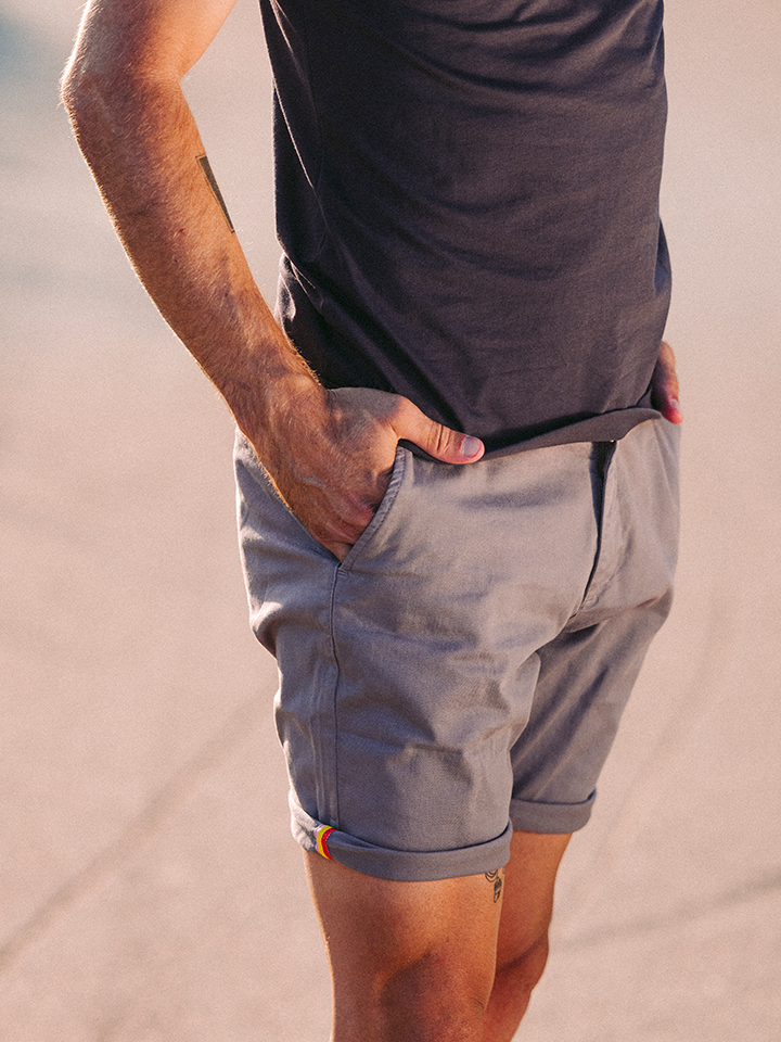 A person stands outdoors wearing La Machine Cycle Club’s Grand Tours - Rest Day Bermuda shorts in grey, featuring a small rainbow detail on the cuff and a semi-tailored fit. Hands are in pockets; a dark tee completes the relaxed look. Background is blurred.
