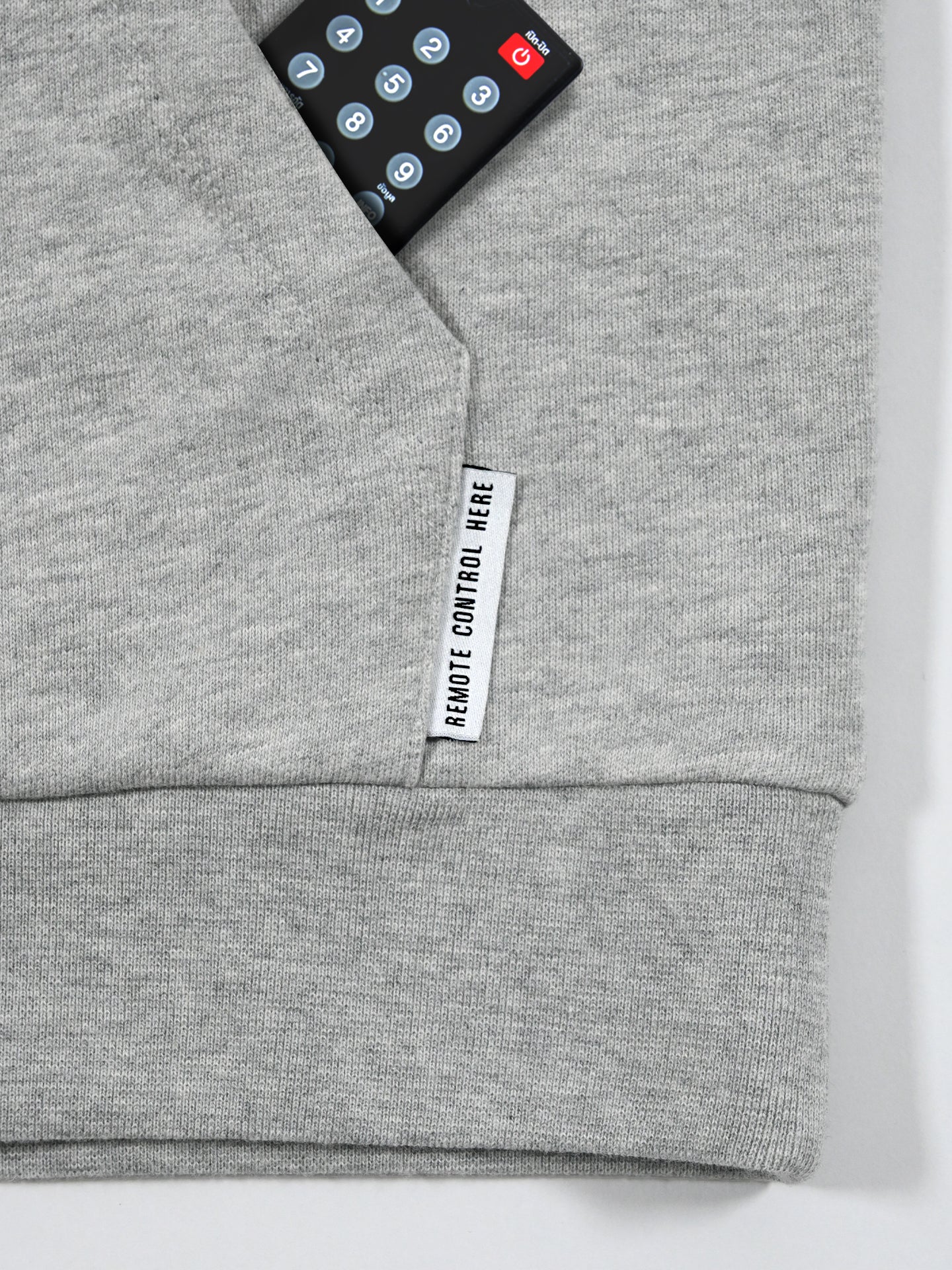 A black remote peeks from a secret pocket on the Beyond the bike - Hoodie - Melange Grey by La Machine Cycle Club, featuring a tag reading REMOTE CONTROL HERE. This unisex hoodie blends style with hidden convenience.