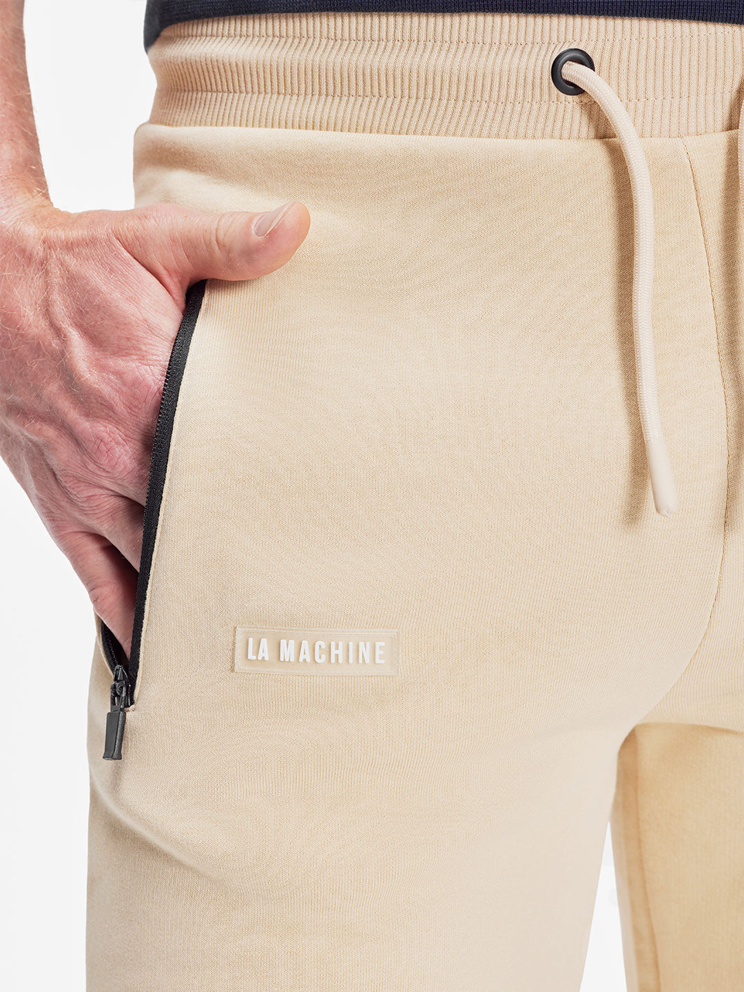 A close-up of a person wearing La Machine Cycle Clubs Beyond the Bike - Sweatpants - Gravel Grey, featuring black zippered pockets, a white drawstring, and an LA MACHINE label. Their left hand is partially in the pocket.
