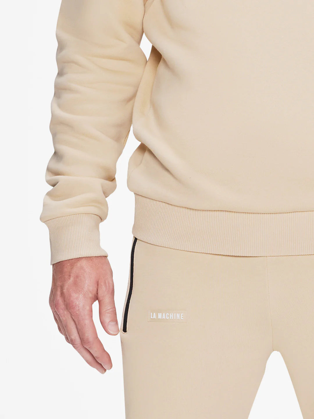 A person wears the La Machine Cycle Club Beyond the bike Hoodie + Sweatpants bundle in Gravel Grey, with LA MACHINE on the pants, shown from shoulder to mid-thigh, one hand relaxed by their side.