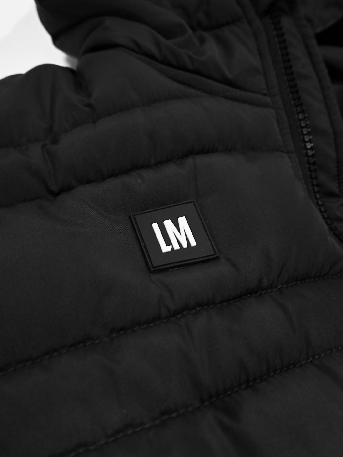 Close-up of the Beyond the bike - Padded Jacket in black by La Machine Cycle Club, featuring quilted padding, a visible zipper, and a square black patch with white LM letters.