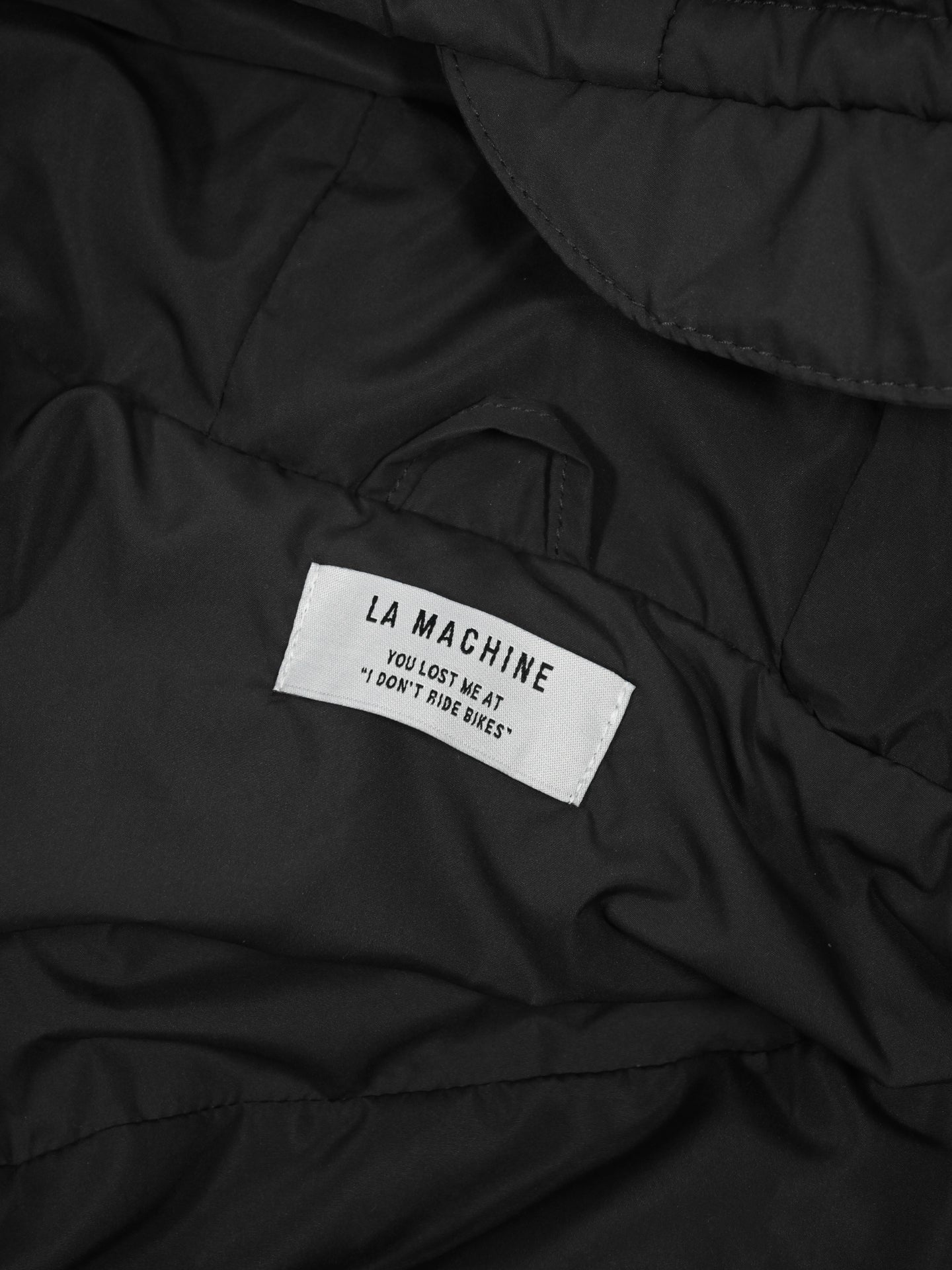 A close-up of the Beyond the bike - Padded Jacket in black by La Machine Cycle Club, featuring a white label with the text: LA MACHINE. YOU LOST ME AT I DONT RIDE BIKES.