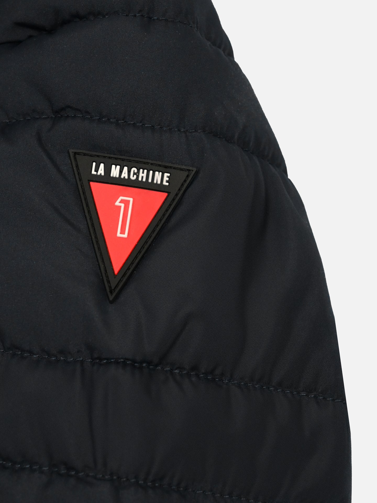 A close-up of the Beyond the bike - Padded Jacket - Black by La Machine Cycle Club, showing a red and black triangular patch with LA MACHINE at the top and the number 1 in the center.