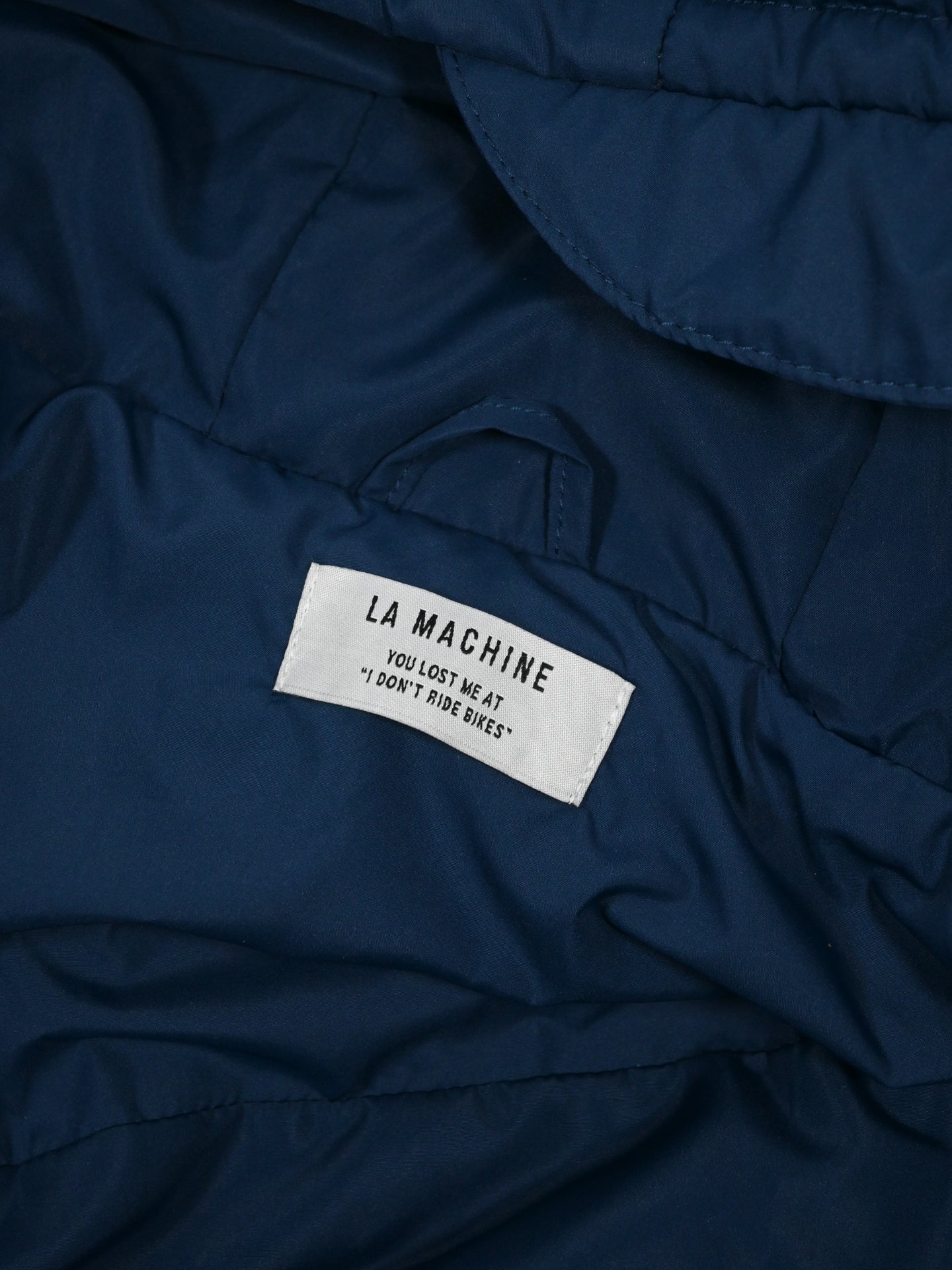 A close-up of the Beyond the bike - Padded Jacket - Blue from La Machine Cycle Club, featuring a white label with the brand name and the phrase: You lost me at I dont ride bikes.
