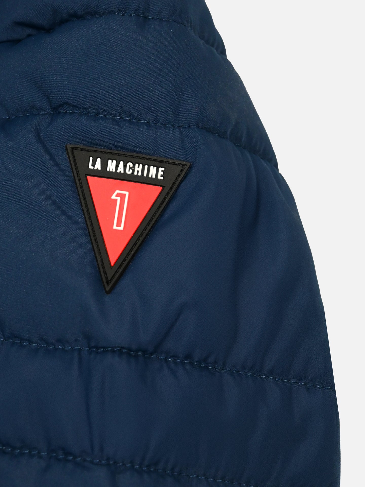 Close-up of the Beyond the bike - Padded Jacket in blue by La Machine Cycle Club, featuring a triangular LA MACHINE patch with a white number 1 on a red background at the top.