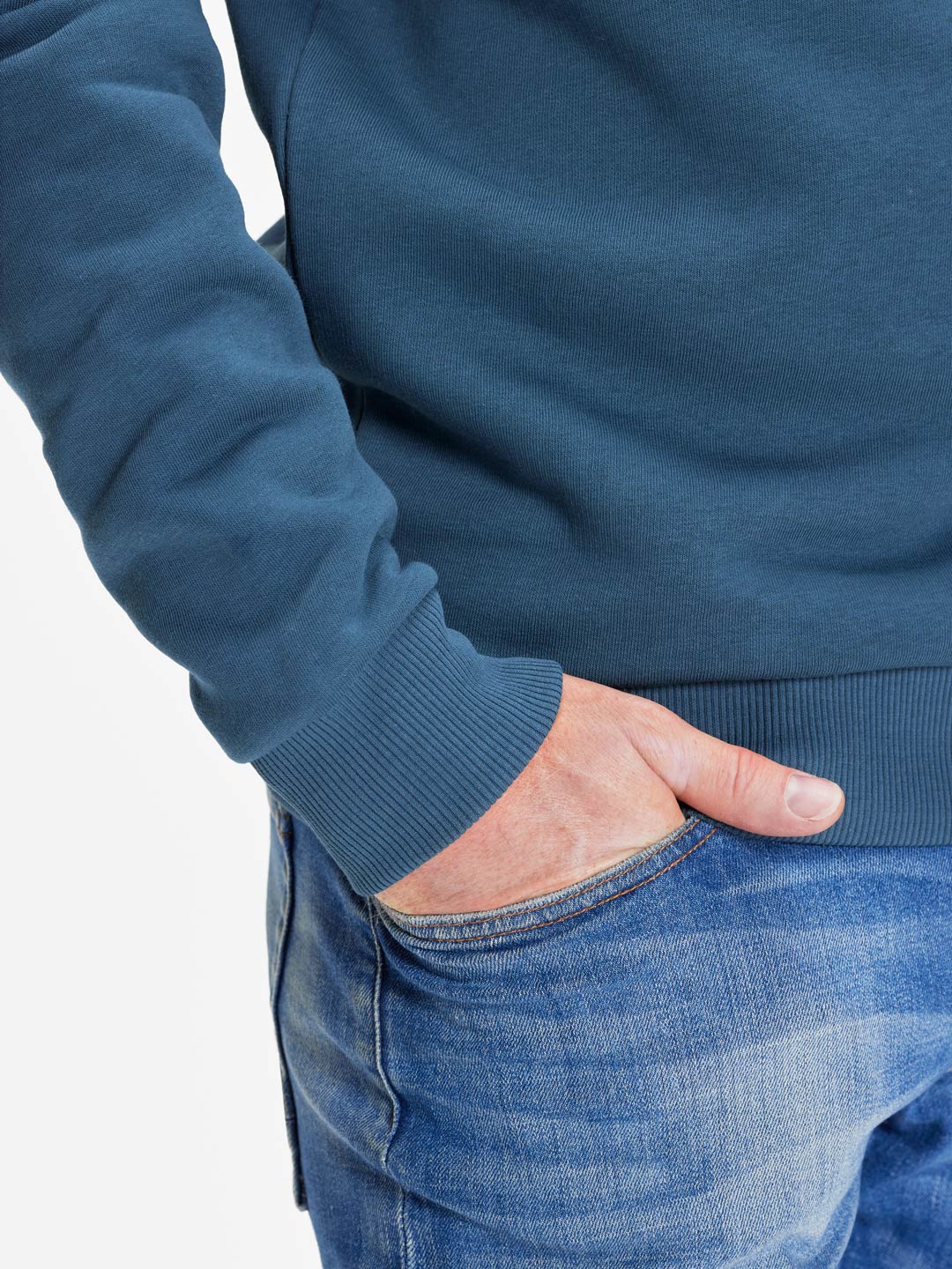 A person wears the La Machine Cycle Club Beyond the Bike - Hoodie in Orion Blue with blue jeans, standing waist down with one hand in their pocket against a white background.