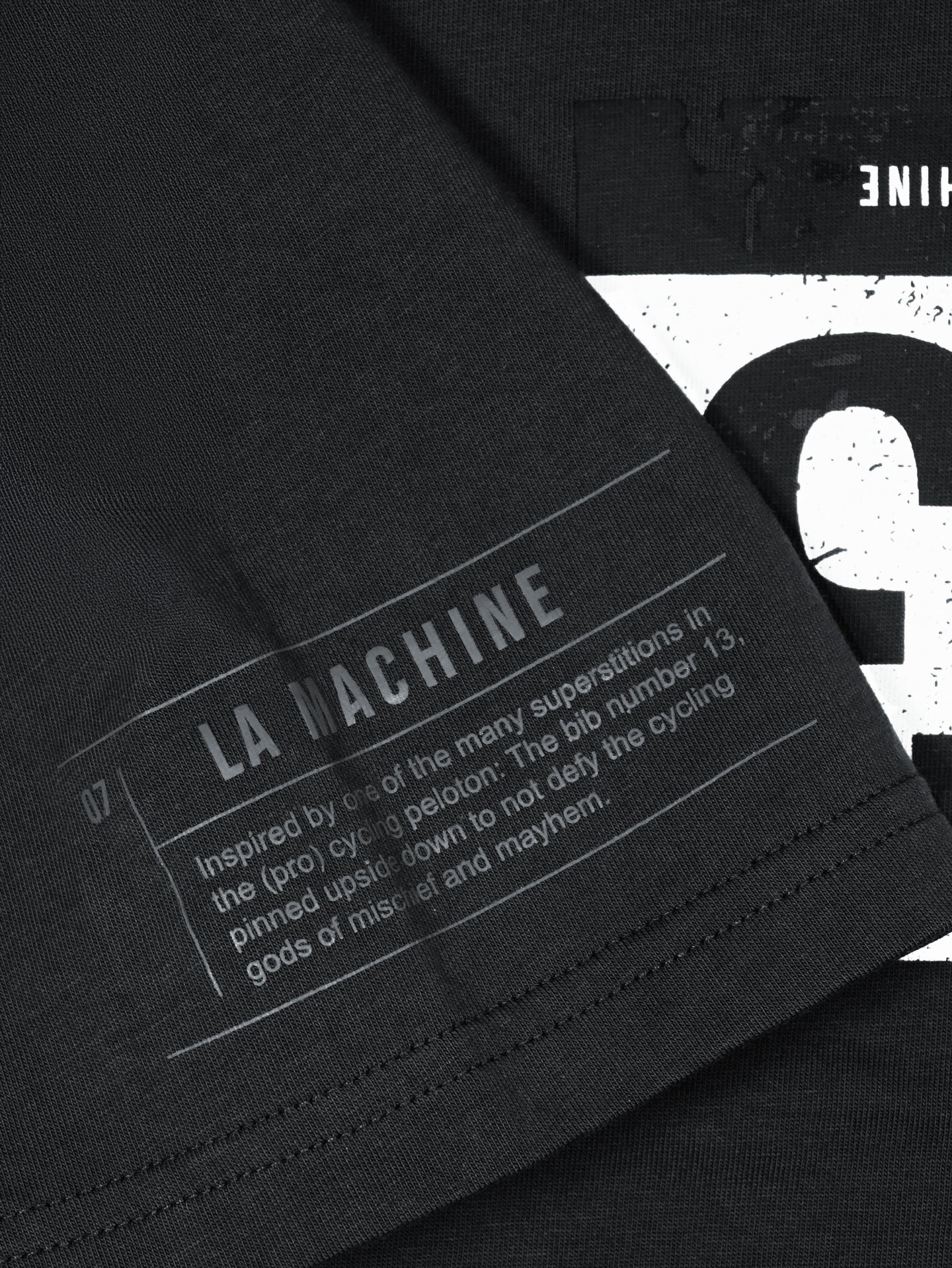 Close-up of a black fabric label on the Bib Number 13 T-shirt in Charcoal Teal by La Machine Cycle Club, featuring white “LA MACHINE” text and a nod to the cycling superstition of pinning bib number 13 upside down for luck.