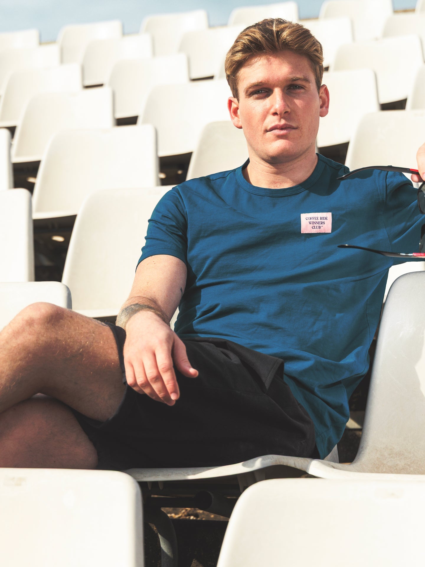 A young man with light brown hair sits relaxed on white stadium seats, wearing the La Machine Cycle Club Coffee Ride Winners Club blue T-shirt and black shorts, looking at the camera with a neutral expression after a casual coffee ride.
