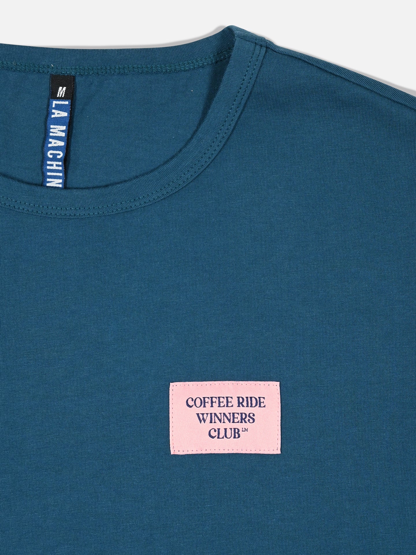 A close-up of the La Machine Cycle Club Coffee Ride Winners Club unisex blue T-shirt, featuring a pink chest patch and black tag at the neckline—ideal for cycling fans.