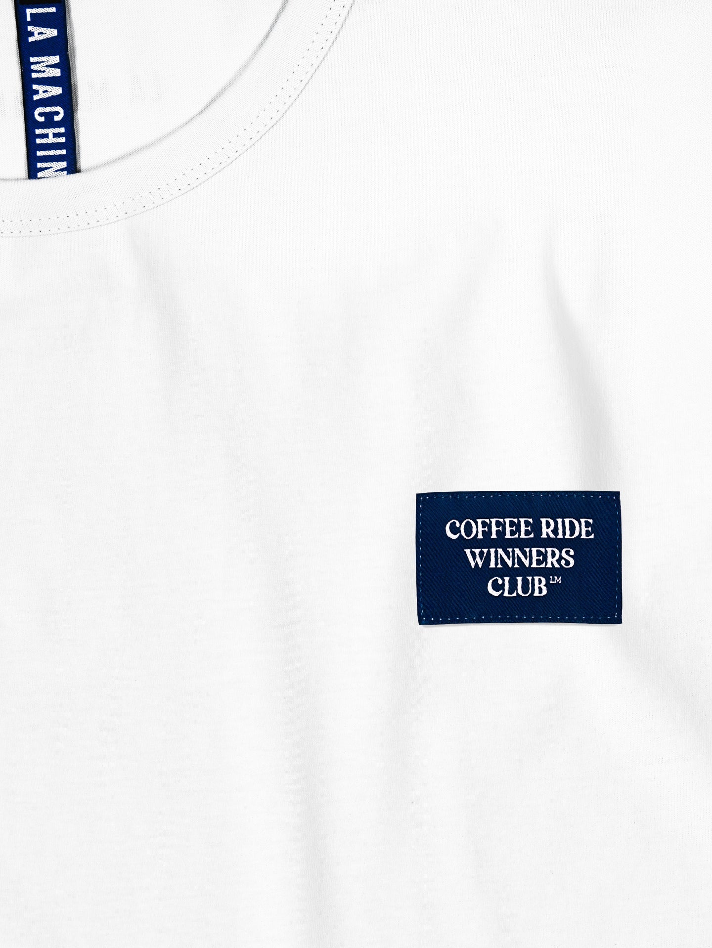 A close-up of the Coffee Ride Winners Club - T-shirt - White by La Machine Cycle Club, featuring a small blue COFFEE RIDE WINNERS CLUB™ patch on the chest and a partially visible blue neck tag at the collar.