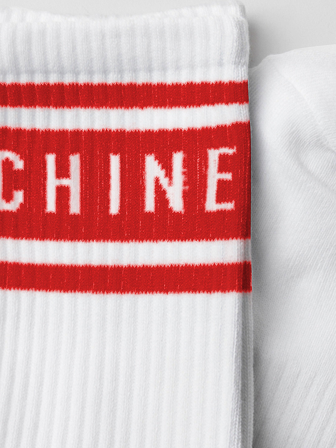 Close-up of La Machine Cycle Club’s La Machine - Crew Socks - Vuelta Red: a white ribbed sock with two Vuelta-red stripes and partial CHINE text, perfect for any cyclist. Another sock is partly visible to the right.