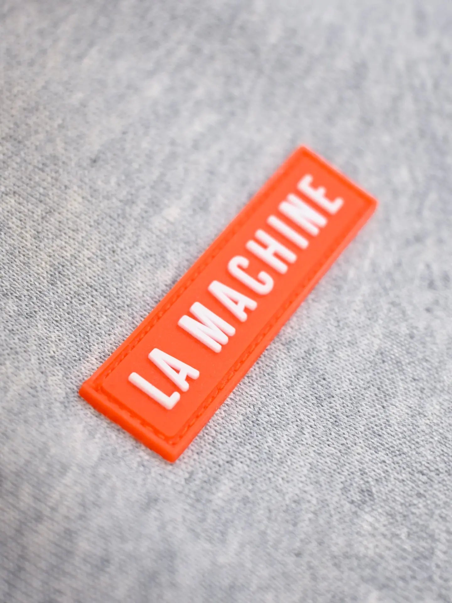 A close-up of the gray De Koersbroek Sweatpants from La Machine Cycle Club, featuring an orange rectangular patch with embossed white LA MACHINE text, showcases their stylish design.