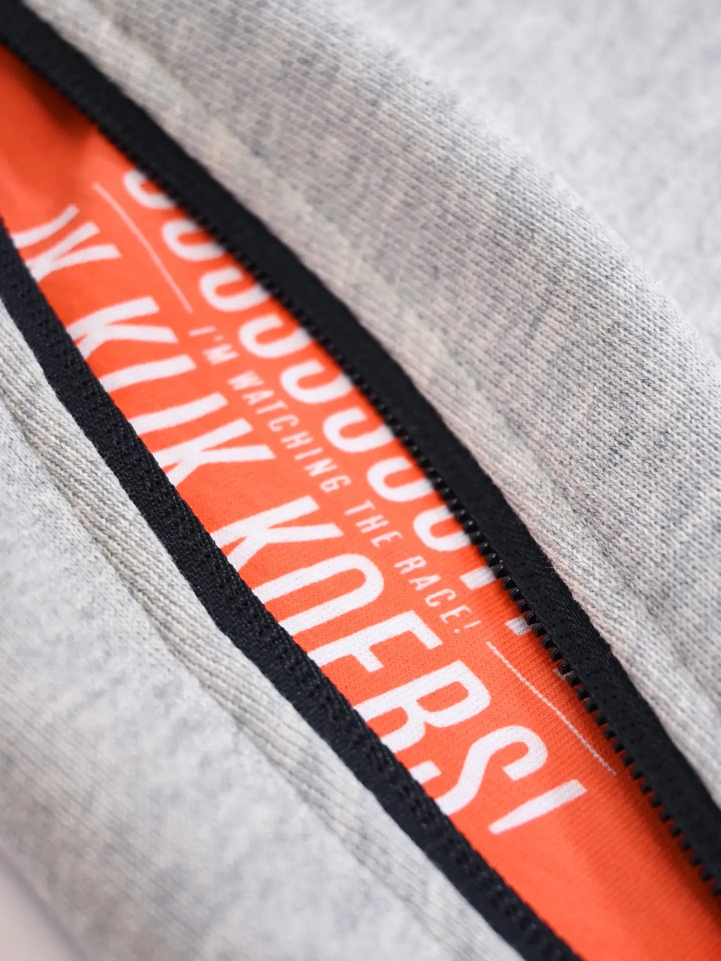A close-up of a partially unzipped light gray hoodie reveals bright orange fabric with white “I’M WATCHING THE RACE! KIJK KOERS!” text—perfect to pair with La Machine Cycle Club’s De Koersbroek - Sweatpants.