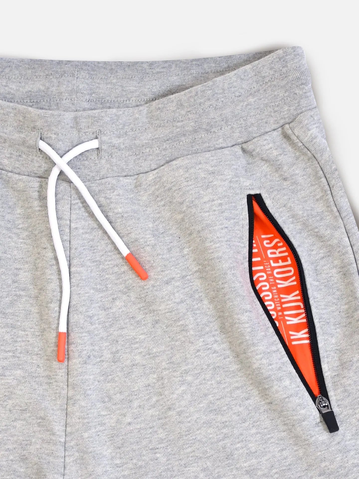 De Koersbroek Sweatpants by La Machine Cycle Club: light gray mens sweatpants with a white drawstring featuring orange tips, a half-open zipped pocket revealing orange lining with white text. Ideal race-viewing wear, displayed on a plain white background.