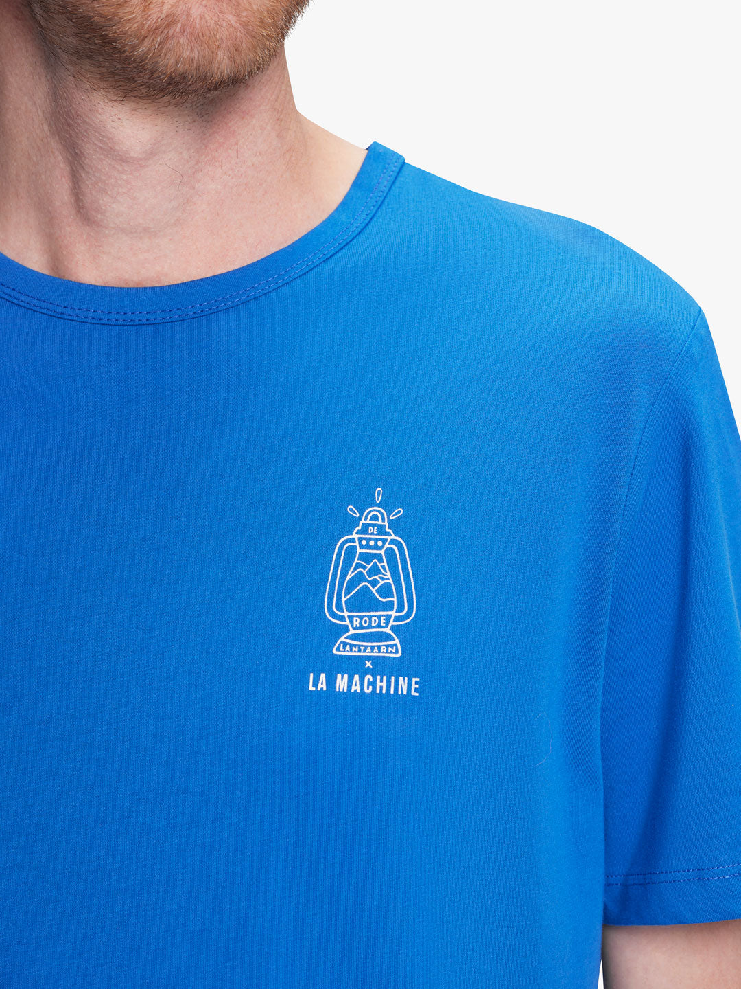 A person wears the De Rode Lantaarn - Das War Einmal t-shirt by La Machine Cycle Club, featuring a bright blue unisex fit with a white machine line drawing and LA MACHINE text—only their lower face and upper torso are visible.