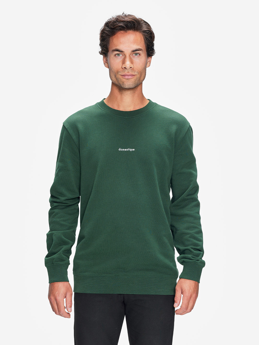 A man with short brown hair faces forward in a La Machine Cycle Club Domestique - Sweatshirt, a dark green mens loose fit top with small white chest text, paired with black pants, set against a plain white background.