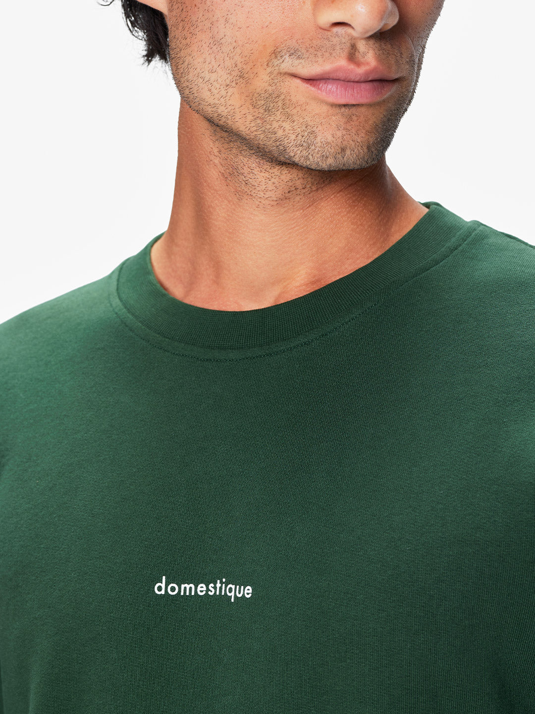 A man wearing the Domestique - Sweatshirt by La Machine Cycle Club in dark green, featuring small white domestique lettering on the chest. Only his lower face, neck, and upper shoulders are visible.