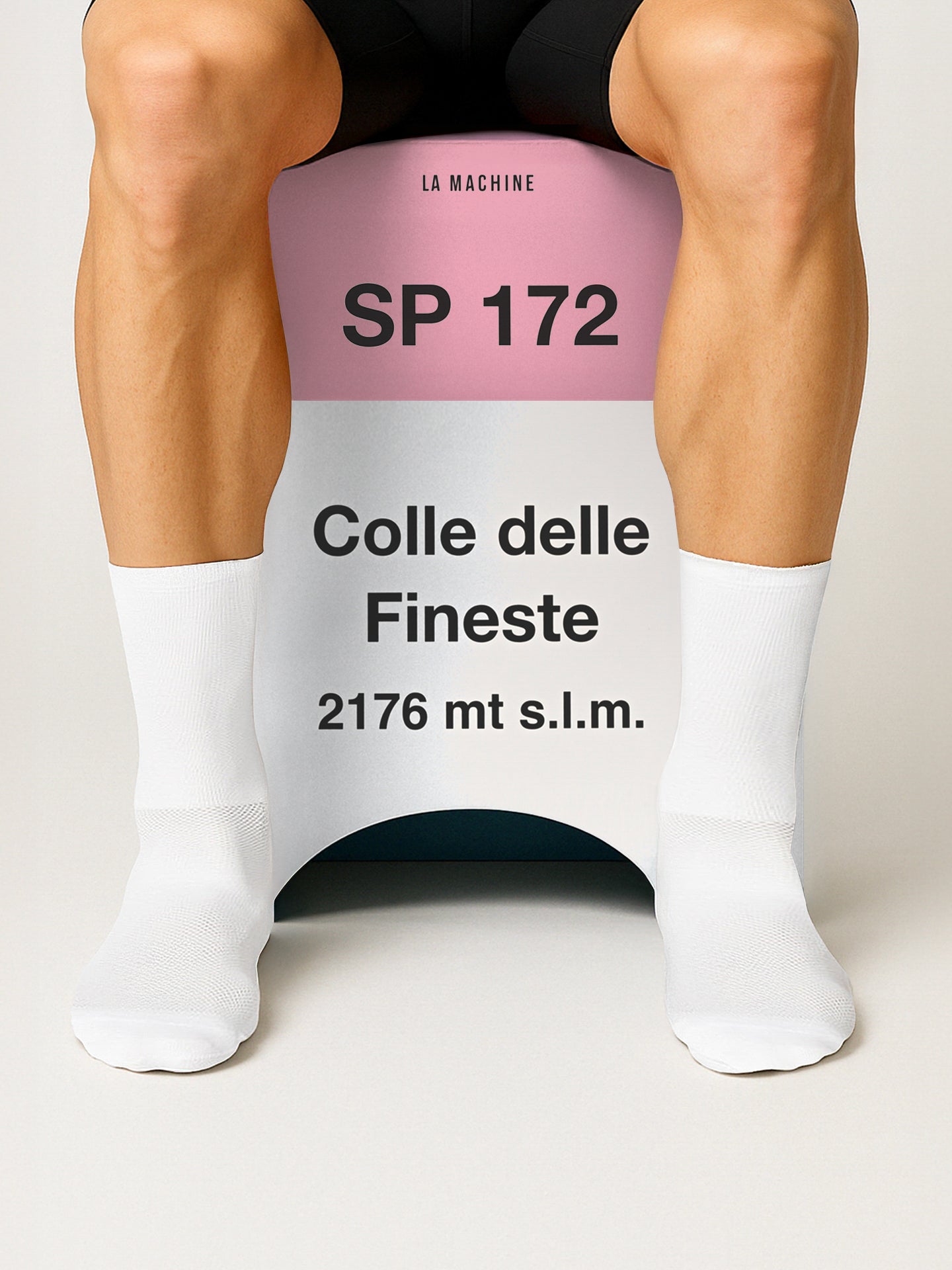 A person in white socks sits on a La Machine Cycle Club Colle delle Finestre - 2-in-1 Stool & Sidetable (Misprint Edition), styled like a road sign, with legs visible mid-thigh down for a unique cycling memorabilia display.