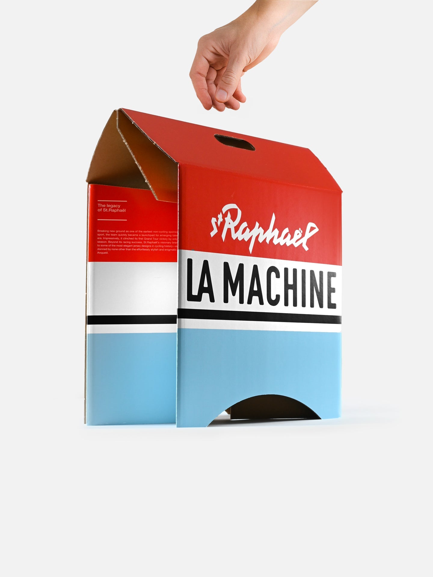 A hand holds a large, red, white, and blue box with a handle, styled like a vintage racing jersey. The box showcases St. Raphaël - 2-in-1 Stool & Sidetable from La Machine Cycle Club against a plain white background.