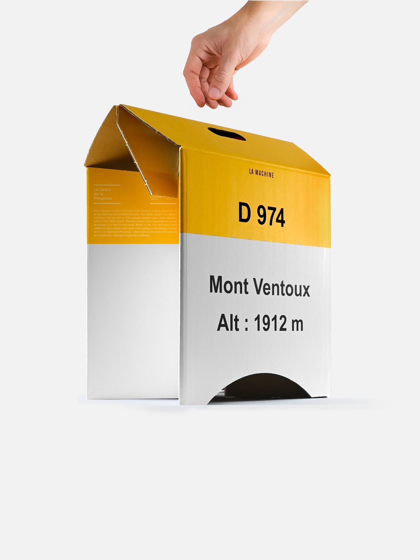 A hand lifts a yellow and white Mont Ventoux - 2-in-1 Stool & Sidetable box by La Machine Cycle Club, with D 974 Mont Ventoux Alt: 1912 m label, open top flap, and semicircular cutout, against a plain white background.