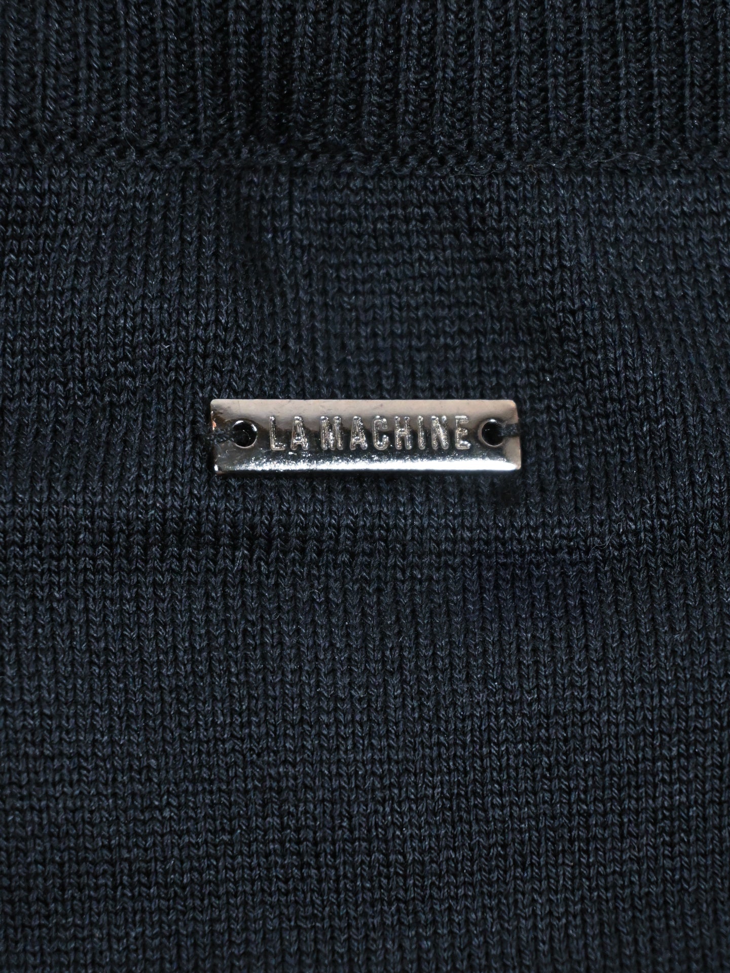 A close-up of the Food of Champions Knitted Pullover by La Machine Cycle Club highlights its refined details, including a small metallic tag with the brand name.
