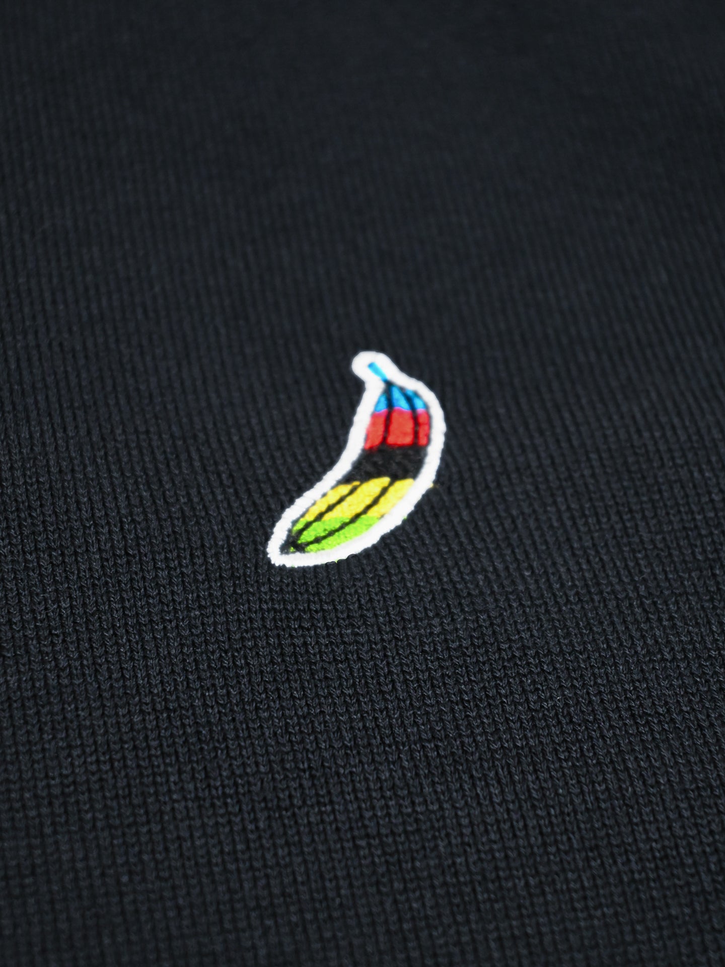 A close-up of black fabric from the La Machine Cycle Club Food of Champions Knitted Pullover, showcasing a small embroidered chili pepper in red, blue, green, and yellow at the center.