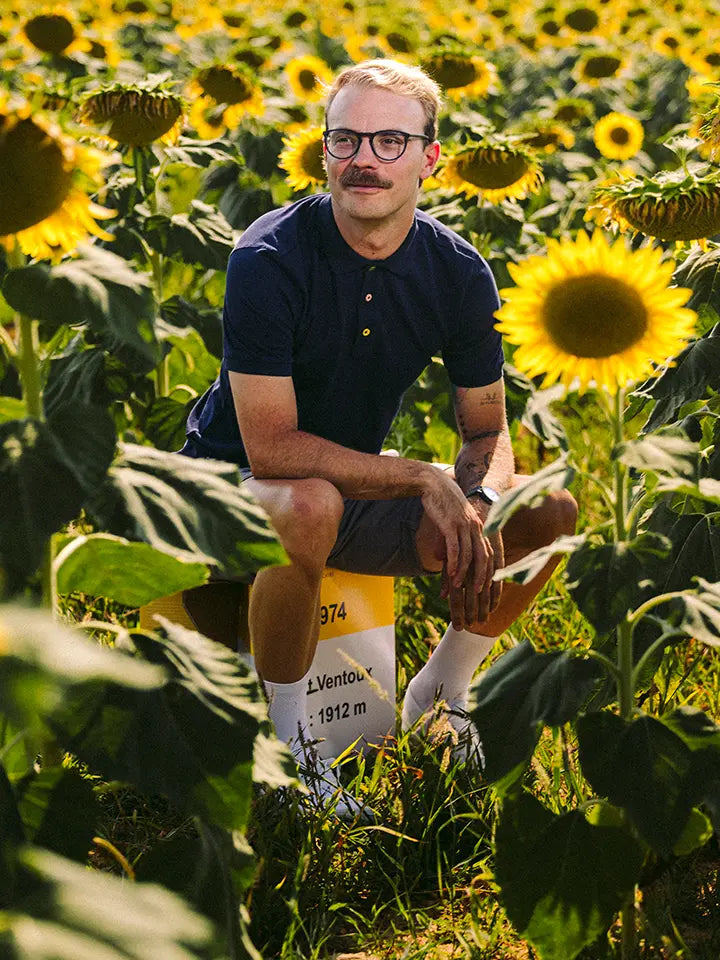 A man with glasses and a mustache sits in a sunflower field, wearing the La Machine Cycle Club Grande Boucle Polo Shirt in navy, paired with shorts and sneakers, surrounded by tall sunflowers under bright sunlight.