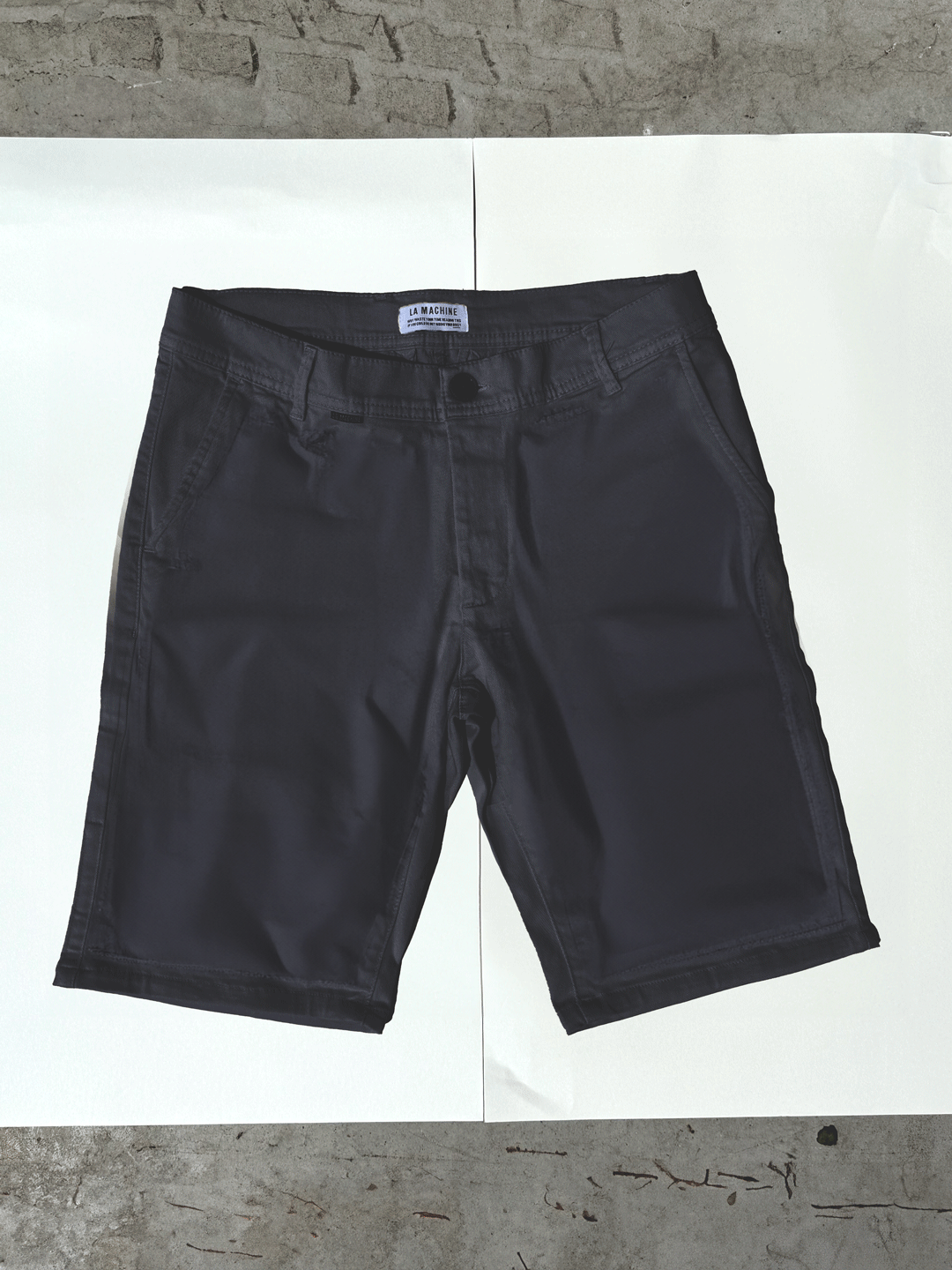A pair of La Machine Cycle Club Grand Tours - Rest Day Bermuda shorts in navy is laid flat on a white surface, with a concrete background partially visible around the edges.