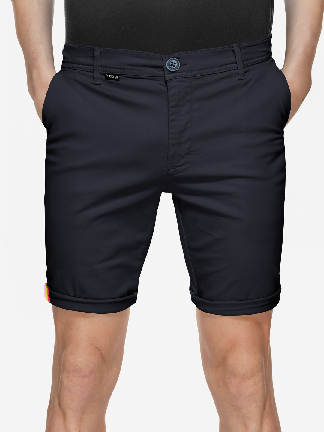 A person wearing La Machine Cycle Club’s Grand Tours - Rest Day Bermuda in navy, featuring a semi-tailored fit, button and zipper closure, side pockets, Grand Tour tape lining, and a small rainbow detail on the lower left hem.