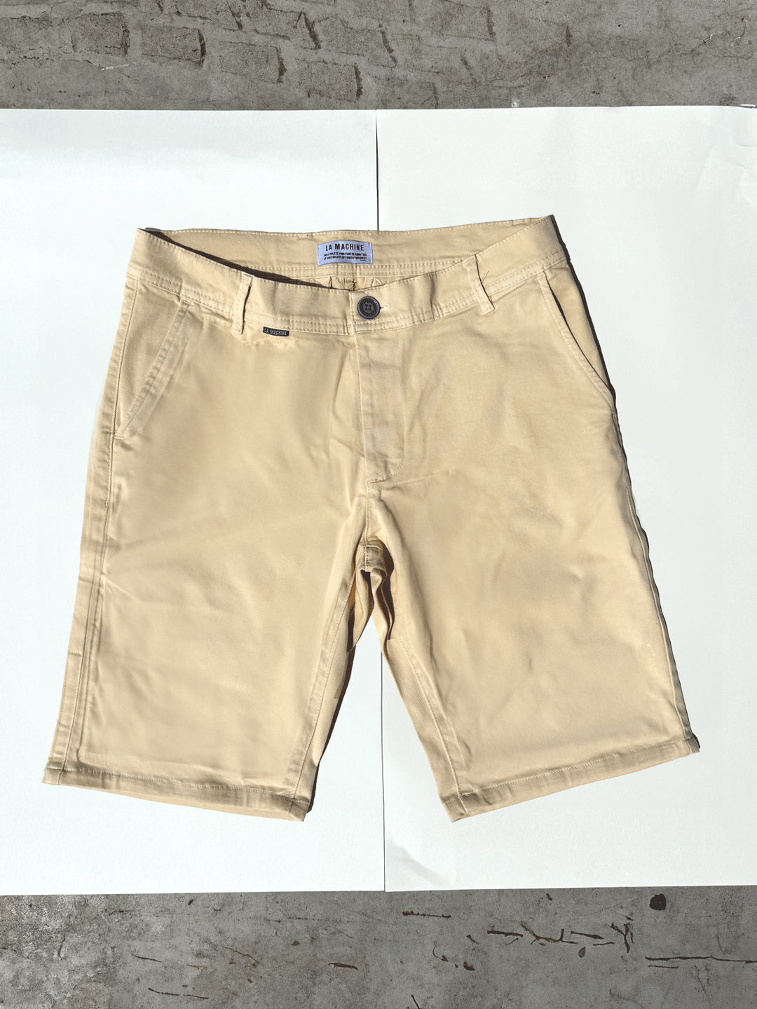 The La Machine Cycle Club Grand Tours - Rest Day Bermuda in Sand, featuring a semi-tailored fit and pockets, is displayed flat on a white surface with a concrete background partially visible at the top and bottom.