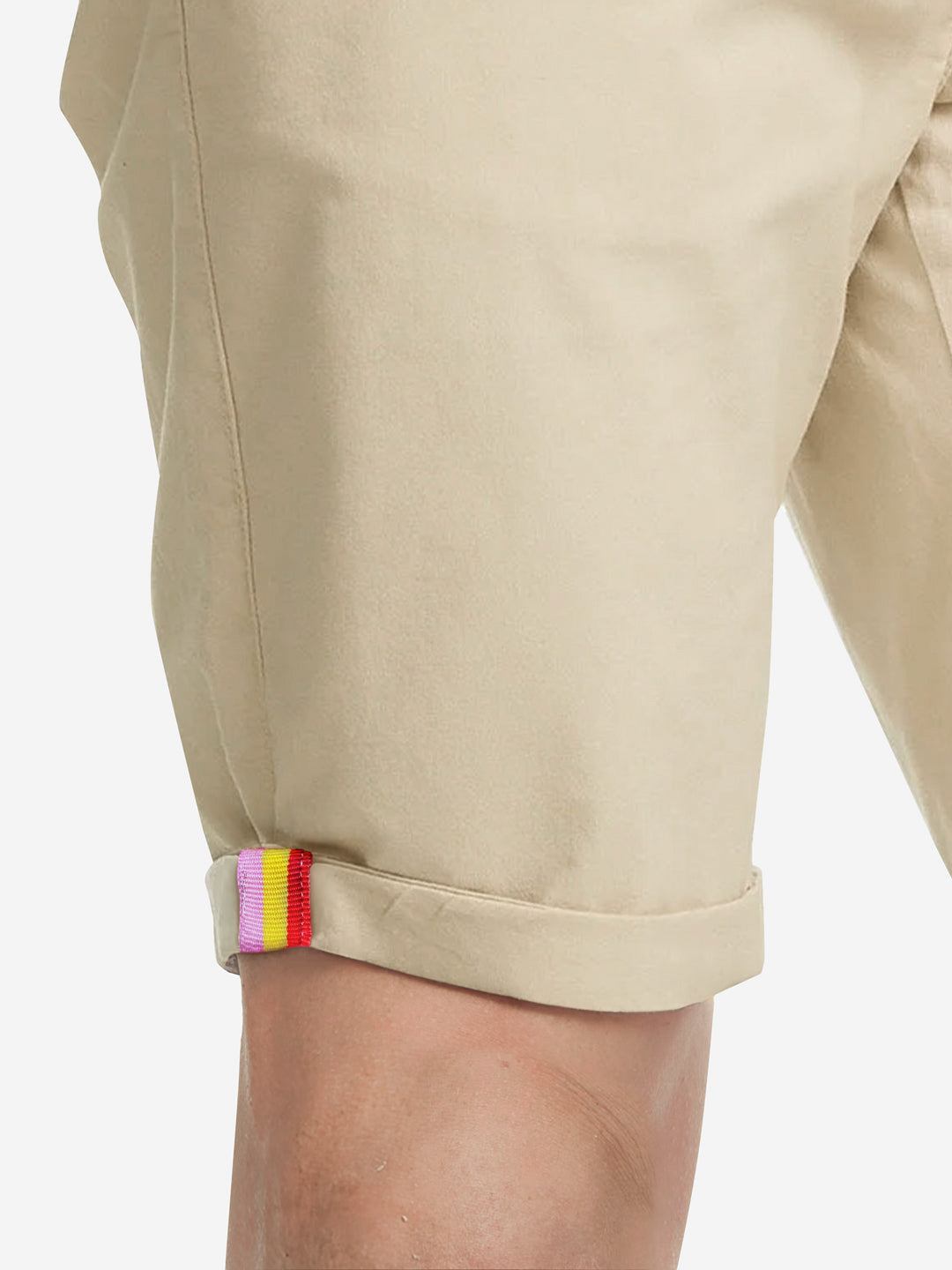 A person wearing the Grand Tours - Rest Day Bermuda in sand by La Machine Cycle Club, semi-tailored mens shorts featuring a Grand Tours colored tape tab in red, yellow, and purple near the rolled-up cuff.