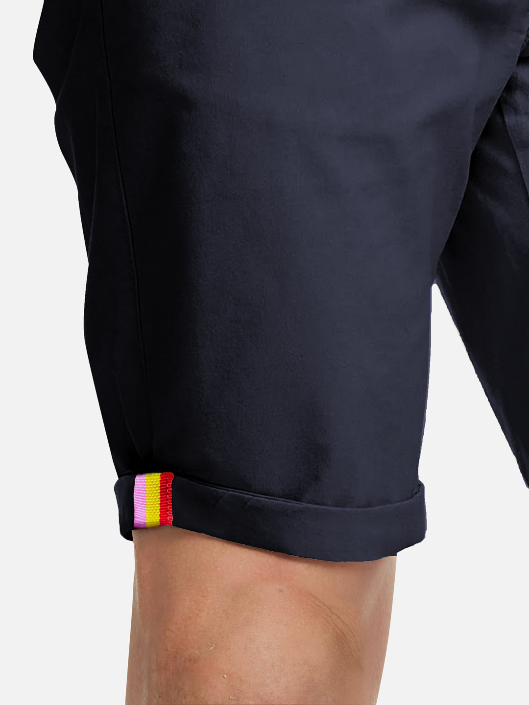 A close-up of a person wearing La Machine Cycle Clubs Grand Tours - Rest Day Bermuda shorts in navy, semi-tailored fit with rolled cuffs and a red, yellow, and white striped tag near the hem. Background is plain white.