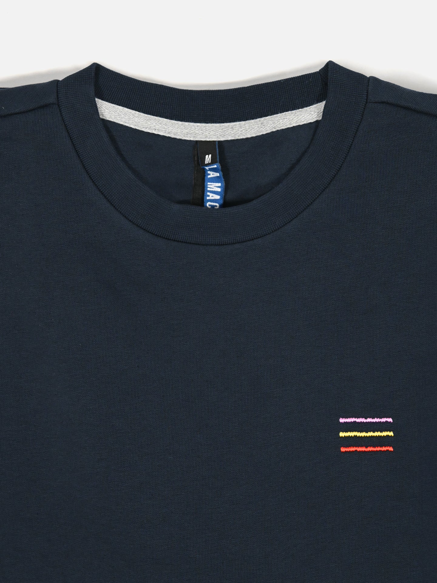 A close-up of La Machine Cycle Clubs Grand Tours III Embroidered Sweatshirt in navy, featuring three Grand Tour-inspired embroidered stripes—yellow, purple, and red—on the left chest. A size M tag is visible inside the collar.